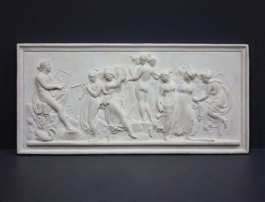 Photo of plaster cast sculpture relief of nude and clothed females (muses) dancing and playing instruments and one male (Apollo) playing a lyre on a dark gray background