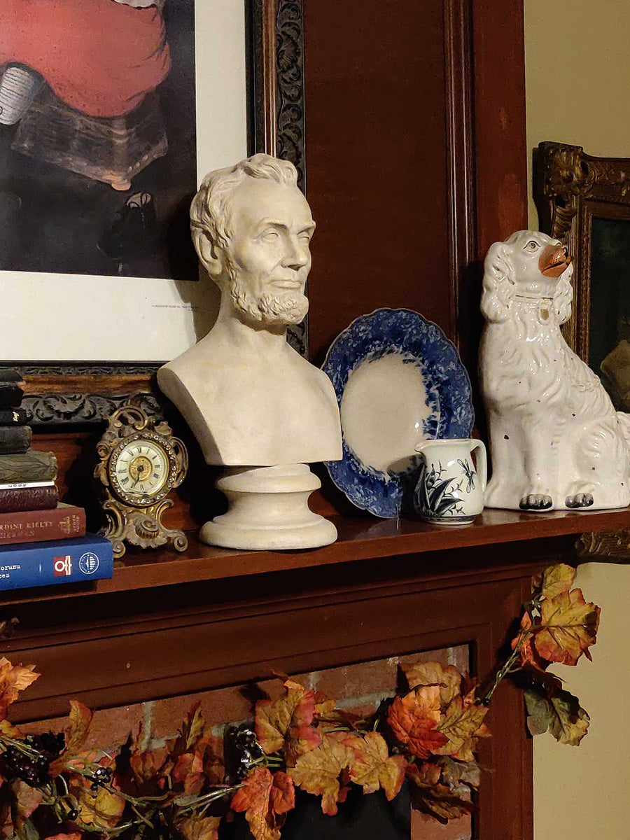 plaster cast bust sculpture of man with beard, namely Lincoln, on dark wood mantel with blue plate, white foo dog, and bronze clock and painting behind