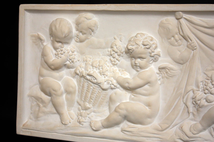 closeup photo of white plaster cast with warm tones of relief sculpture of several cupids talking together with books and baskets of fruit on a black background