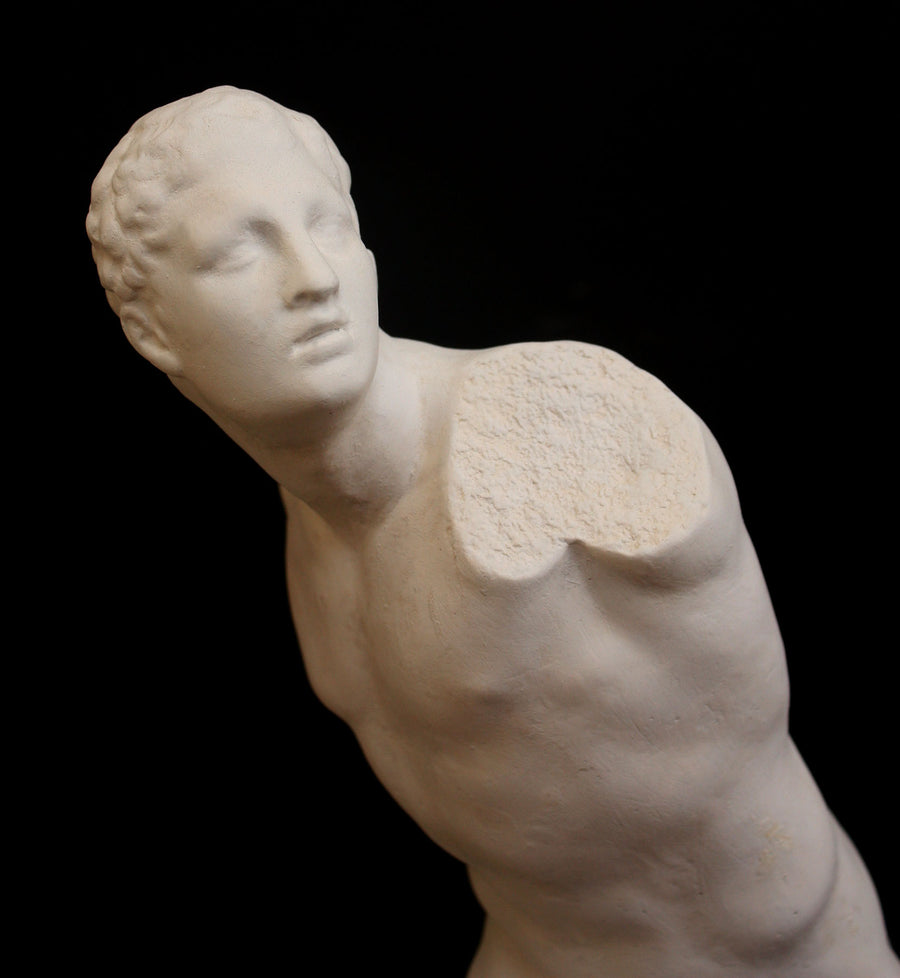 closeup photo of white plaster cast sculpture of male figure in active pose without arms or lower legs on a base against black background