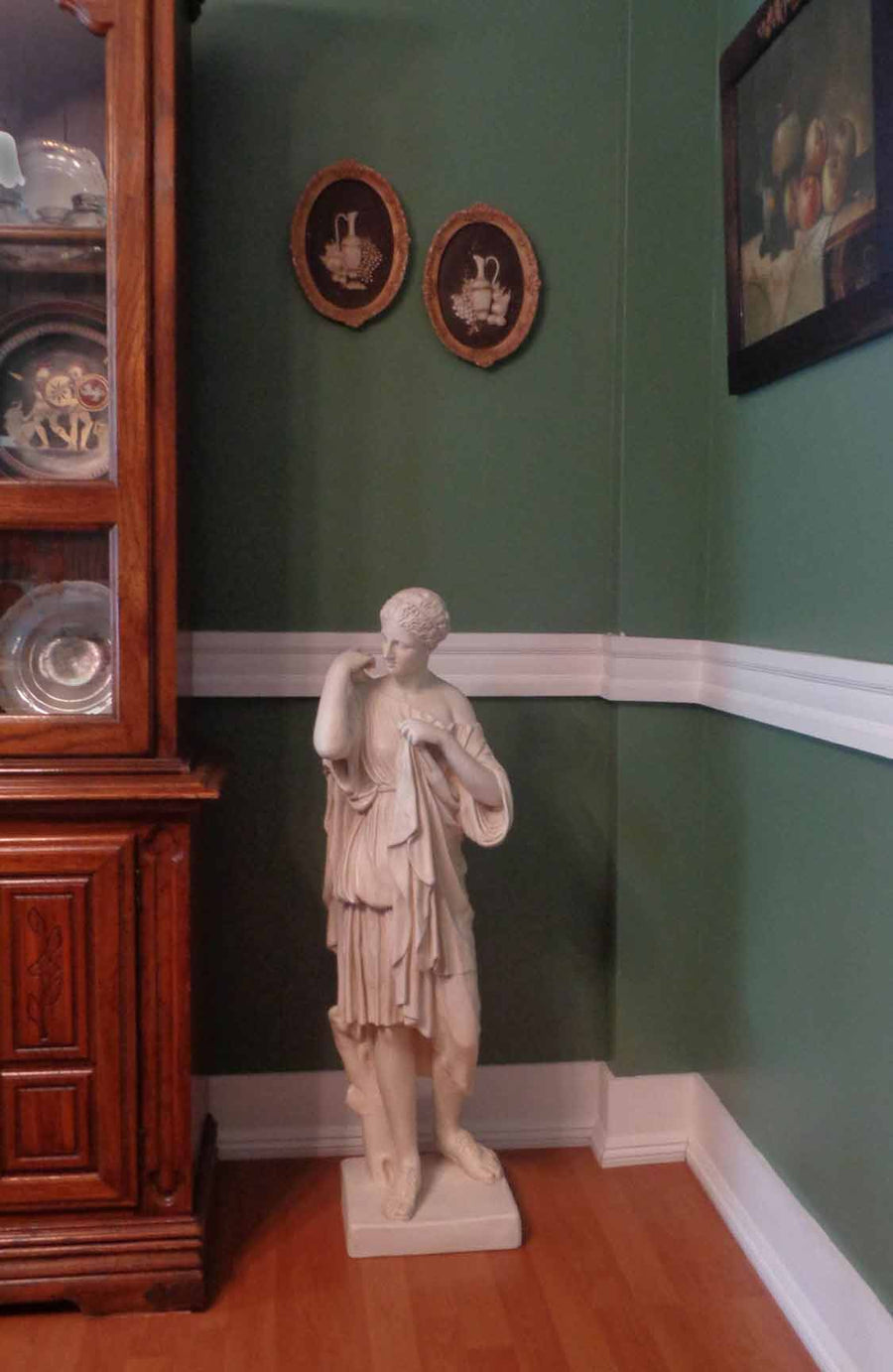 photo of plaster cast of female figure, namely the goddess Diana, with robes on a wood floor in front of a green wall with part of a hutch visible to the left and decorations in hutch and on walls