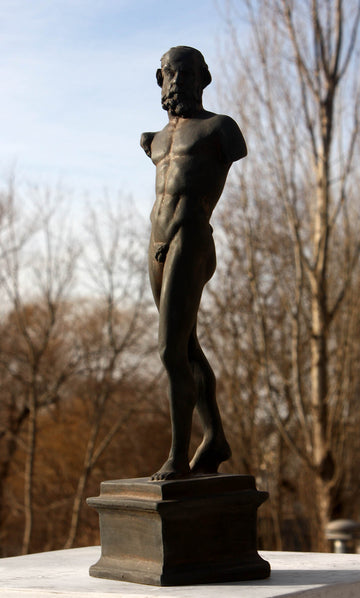 photo of bronze-colored plaster cast sculpture of standing nude satyr figure on square base in front of trees and blue sky