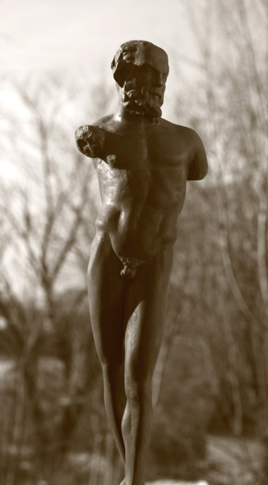 photo detail of bronze-colored plaster cast sculpture of standing nude satyr figure on square base in front of trees
