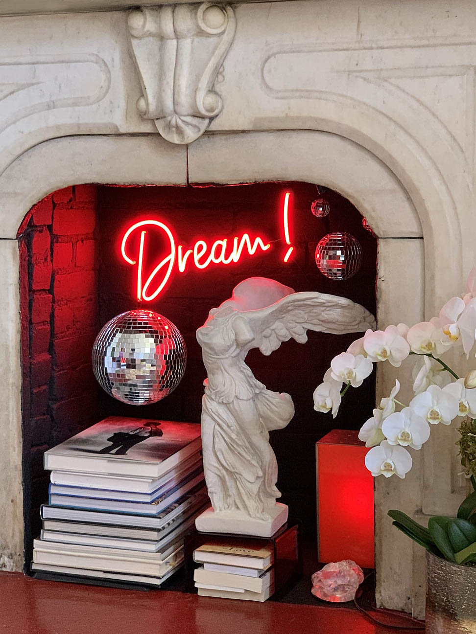 photo of white plaster cast sculpture of winged, headless female figure with flowing drapery in a white, ornate fireplace displayed on a stack of books with another stack of books beside it, disco balls hanging, red neon sign of the word dream hanging, and flowers to the right outside the fireplace