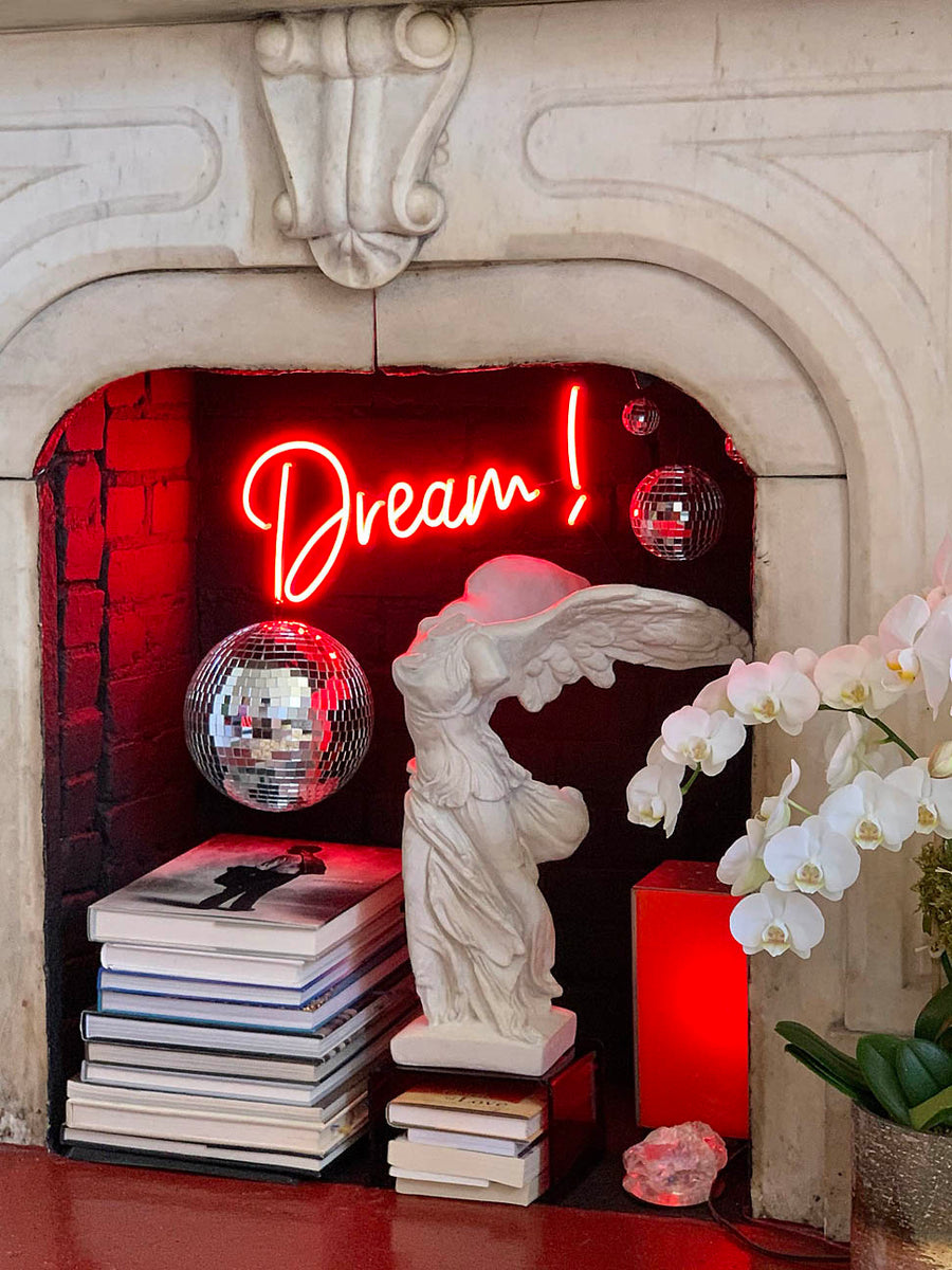 photo of white plaster cast sculpture of winged, headless female figure with flowing drapery in a white, ornate fireplace displayed on a stack of books with another stack of books beside it, disco balls hanging, red neon sign of the word dream hanging, and flowers to the right outside the fireplace