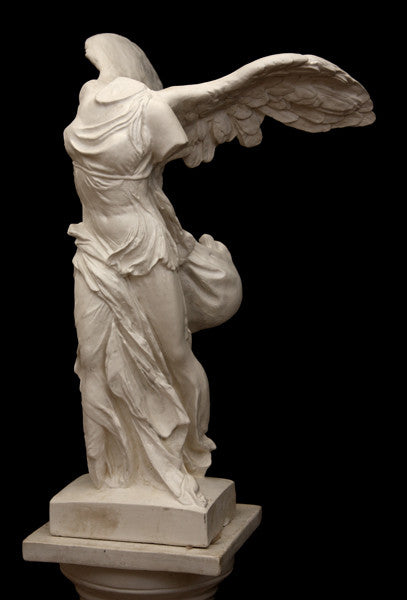photo of plaster cast sculpture of winged, headless female figure with flowing drapery on top of a Doric-column pedestal on a black background