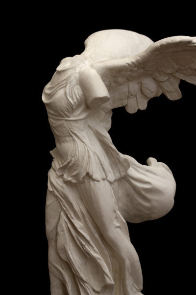 photo of plaster cast sculpture of winged, headless female figure with flowing drapery on a black background