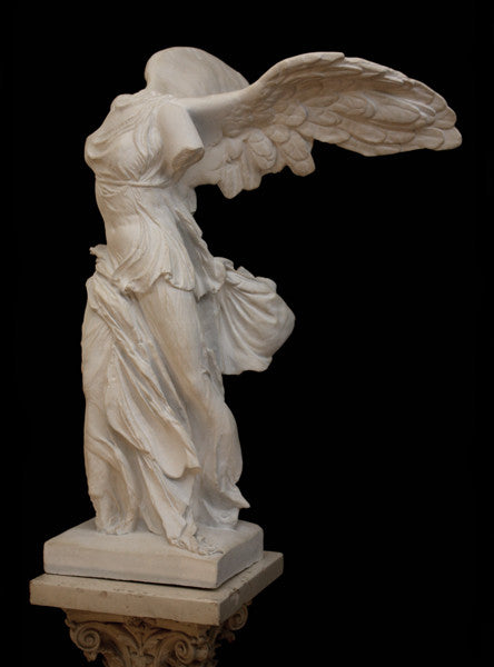 photo of plaster cast sculpture of winged, headless female figure with flowing drapery on top of an ornate, floral pedestal on a black background