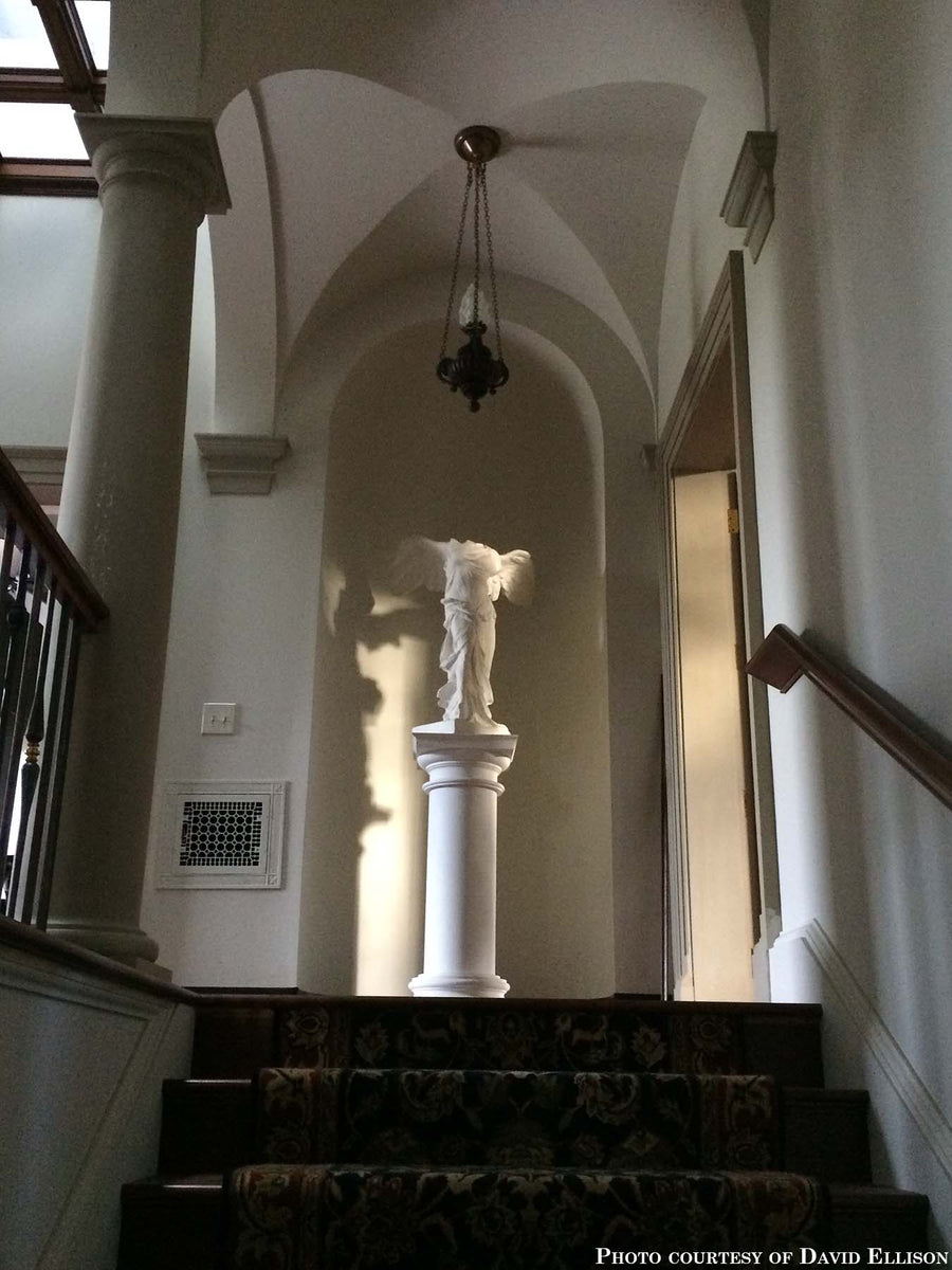 photo of plaster cast sculpture of winged, headless female figure with flowing drapery on pedestal atop a carpeted staircase in a niche in a white-walled room