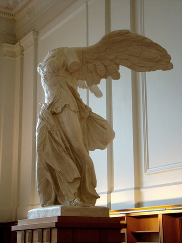photo of plaster cast sculpture of winged, headless female figure with flowing drapery on top of a ribbed, square, wooden pedestal with white paneled walls behind