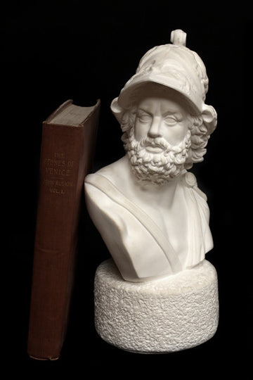 Photo with black background of plaster cast sculpture of male bust with curly hair and beard and helmet and a red book beside it