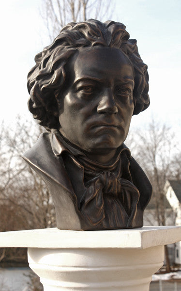 photo of bronze-colored plaster cast sculpture bust of man, namely Beethoven, with neckerchief on pedestal with sky and trees in background
