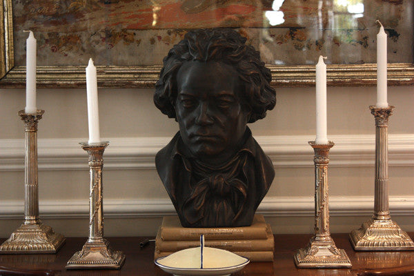 photo of bronze-colored plaster cast sculpture bust of man, namely Beethoven, with neckerchief on dark wood table with candlesticks