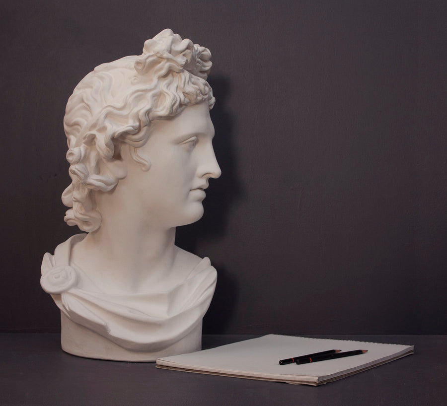 photo of white plaster cast sculpture bust of man, namely the god Apollo, with hair piled high in the front and a broach near his neck holding robes and a sketchbook and pencils beside it on a gray background