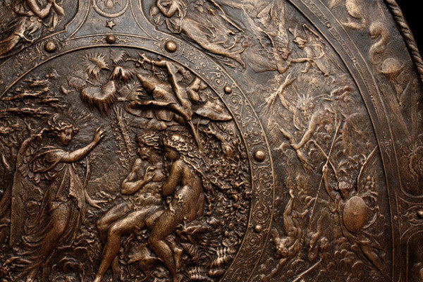 detail photo of bronze-colored plaster reproduction of a shield with intricate relief sculpture set against black background