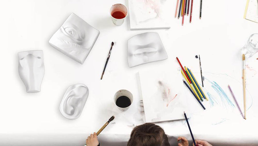 Photo of the top of a white work space with the head of a child drawing with a collection of plaster sculptures, paints, brushes, and paper
