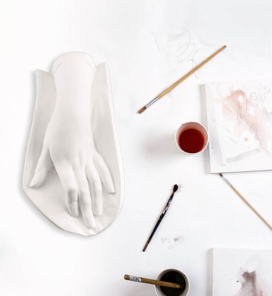 Photo of a plaster cast of a female hand on a white table with paintbrushes, paints, and paper