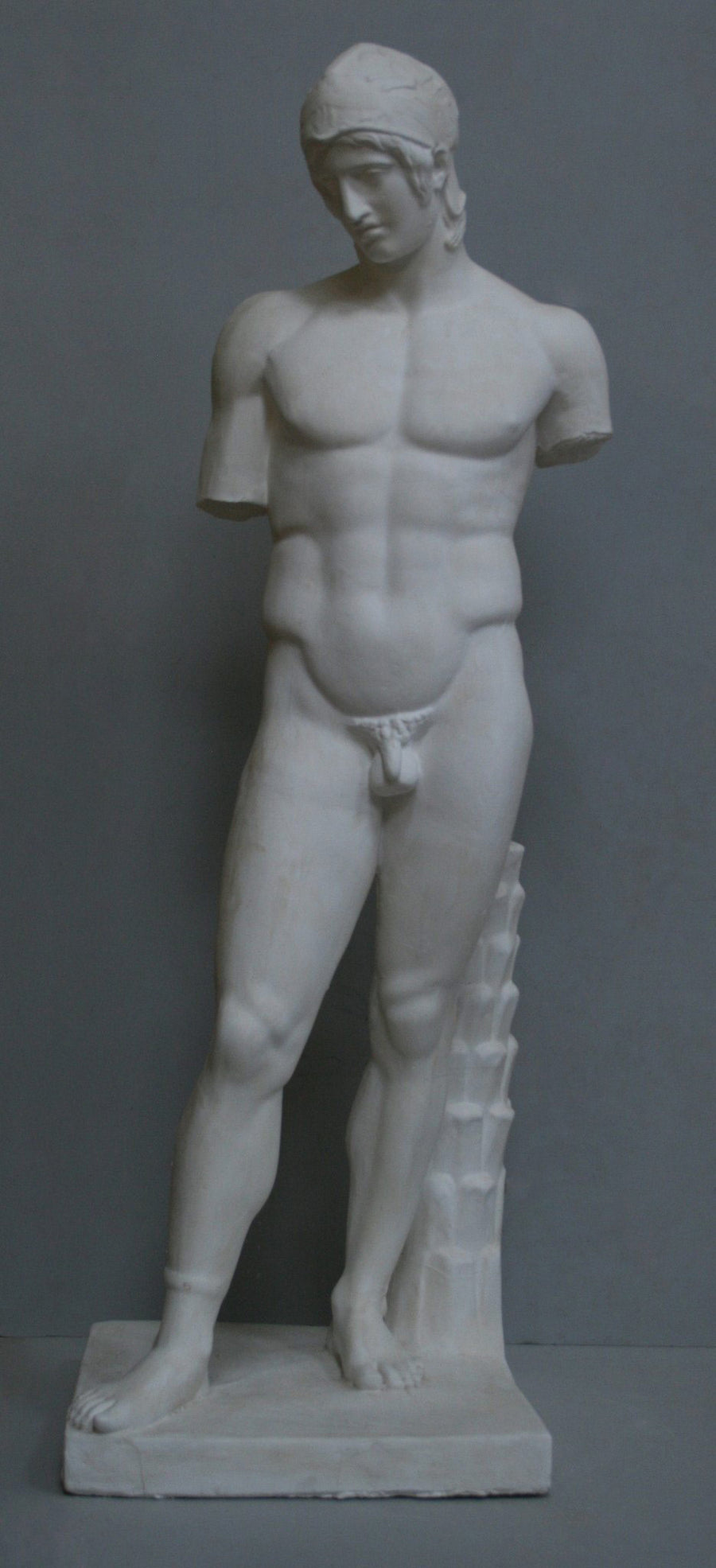 photo of plaster cast of sculpture of nude male, namely the god Ares, without arms and wearing a helmet with a supporting tree trunk, on a gray background