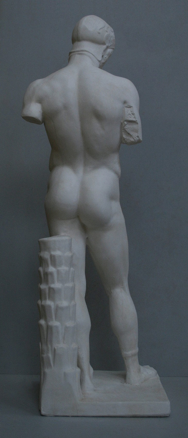photo of back of plaster cast of sculpture of nude male, namely the god Ares, without arms and wearing a helmet with a supporting tree trunk, on a gray background