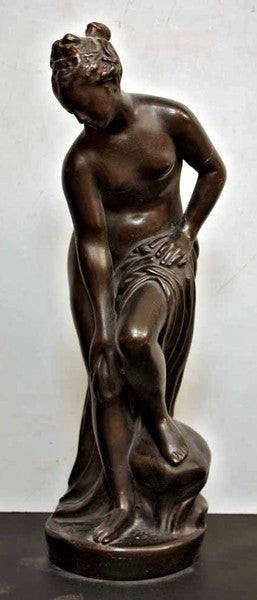photo of bronze-colored plaster cast of sculpture of partially nude female, namely Venus, leaning forward to wash her left leg, placed on a dark table against white background
