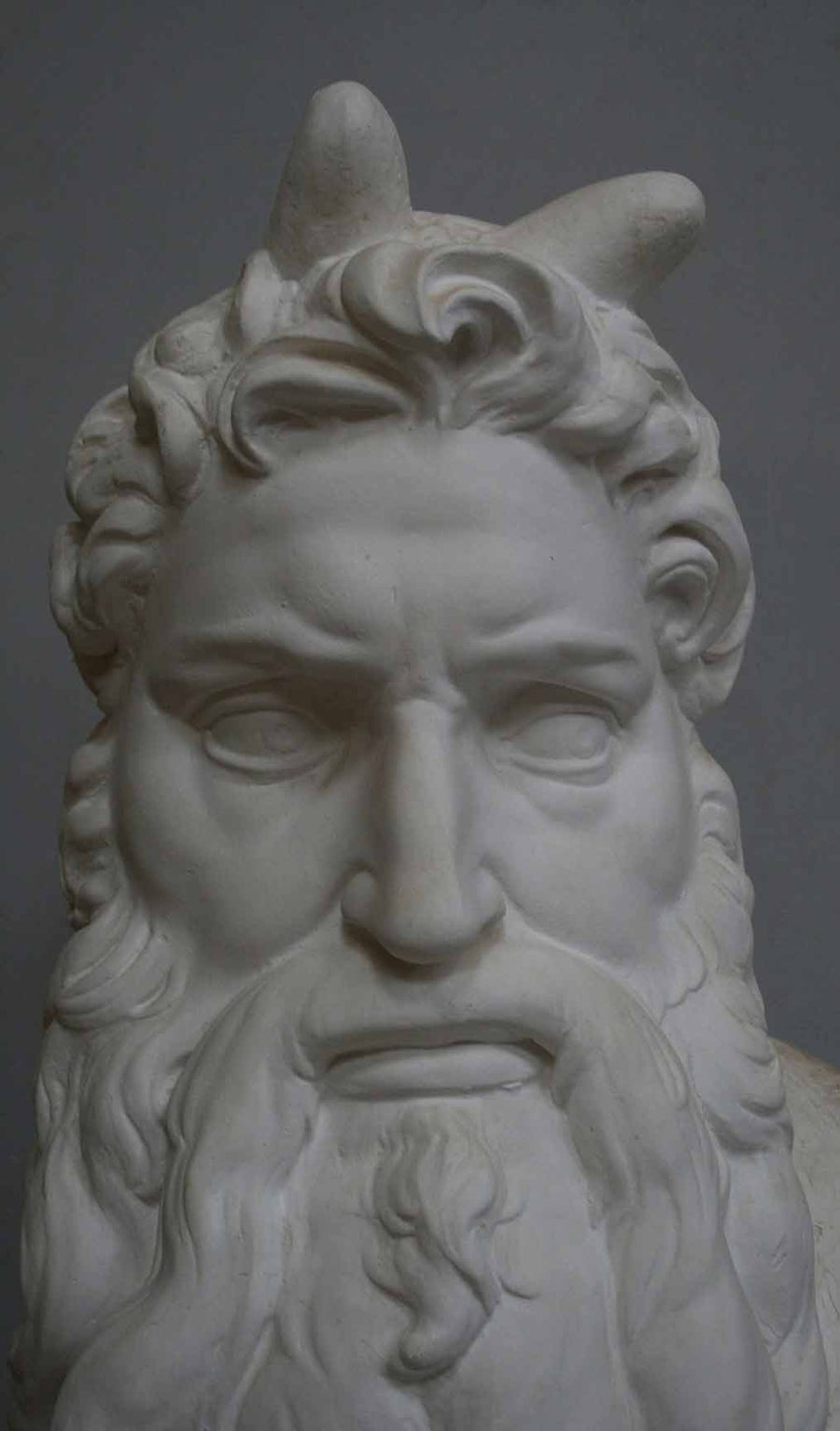 closeup photo of plaster cast bust of man, namely Moses, with curly hair and so-called horns and long beard on gray background