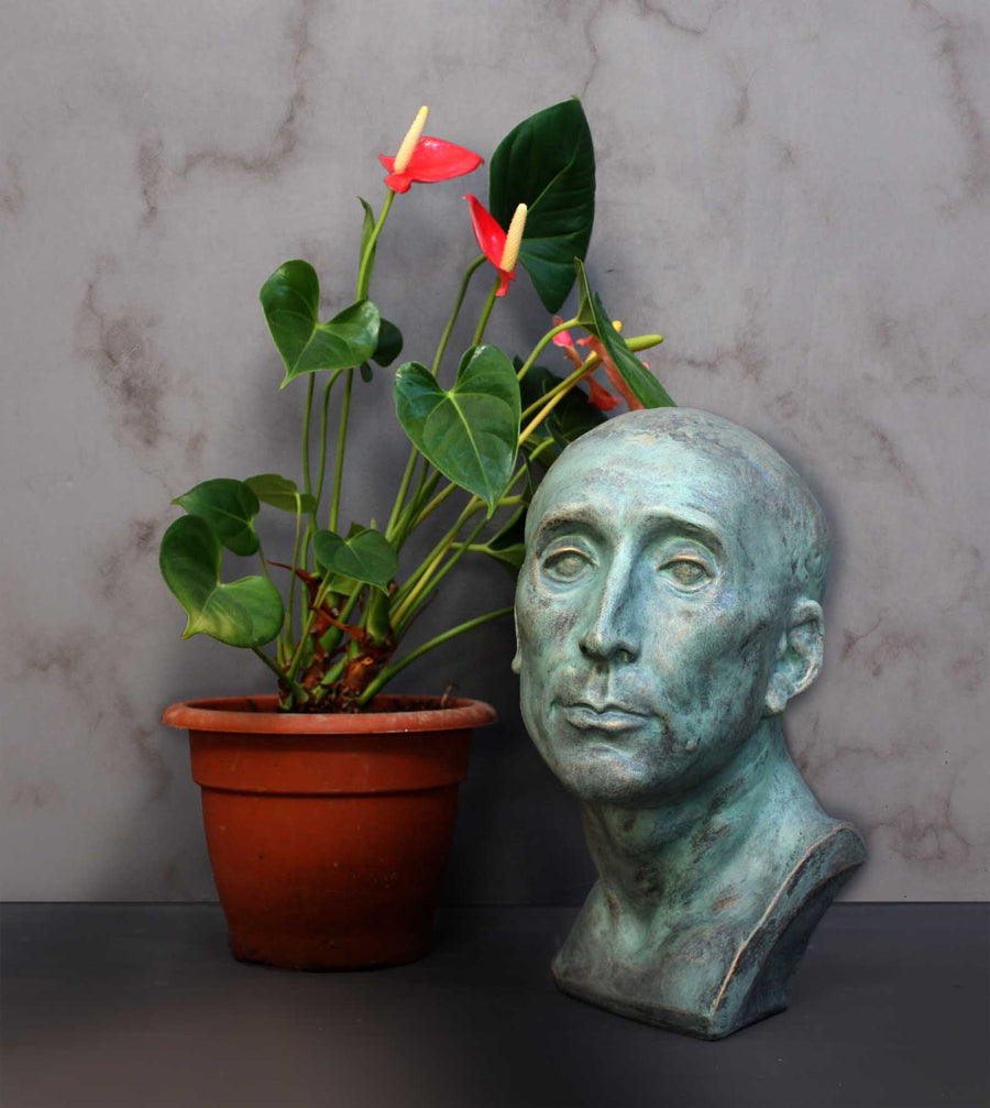 photo of plaster cast sculpture bust of man, namely Niccolo da Uzzano, in verdigris patina next to plant with pink flowers in brown pot in front of a white marble wall and on a gray surface