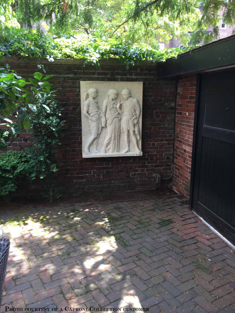 photo of plaster cast of ancient relief with three figures, namely Hades, Eurydice, and Orpheus, on a brick wall outdoors with trees behind, brick paving, and black door to the right