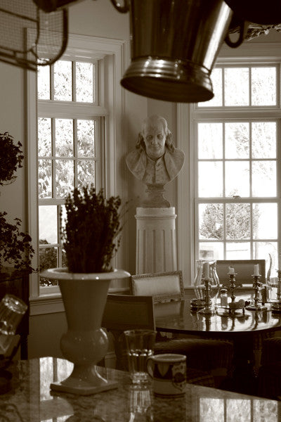 Sepia-toned photo of male bust sculpture on tall pedestal in between two windows in a dining room