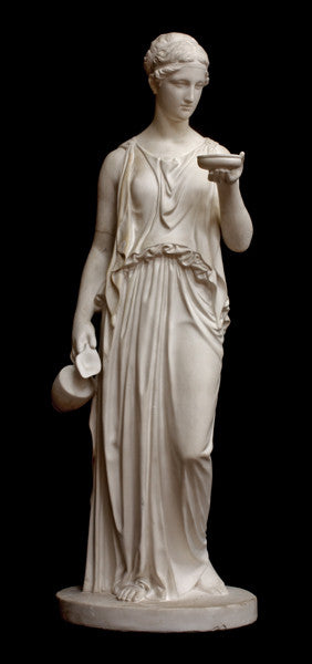 photo with black background of plaster cast sculpture of goddess Hebe in robes standing and holding a jug in right hand at her side and a bowl in left hand raised in front of her