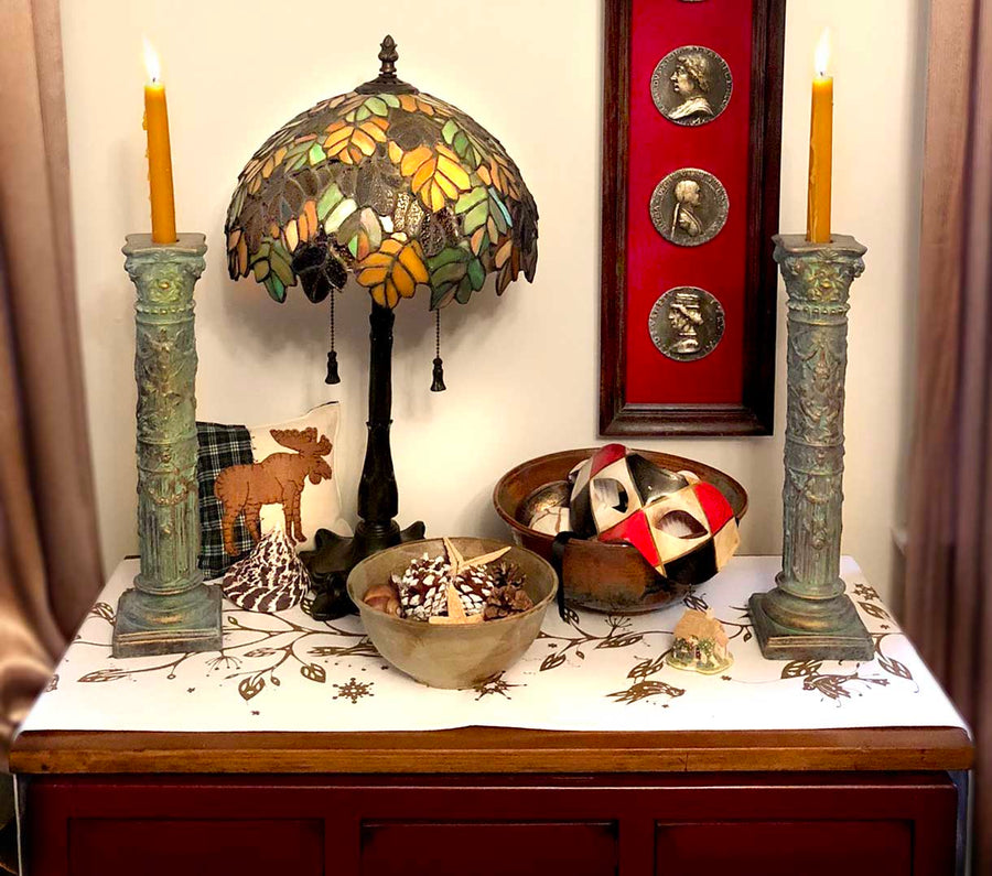 photo of small table with patterned, white tablecloth, two verdigris-color plaster candlesticks with lit orange candles, colorful glass lamp, decorative bowls with pine cones and a Venetian mask, pillow with moose image, and on off-white wall behind, a ta