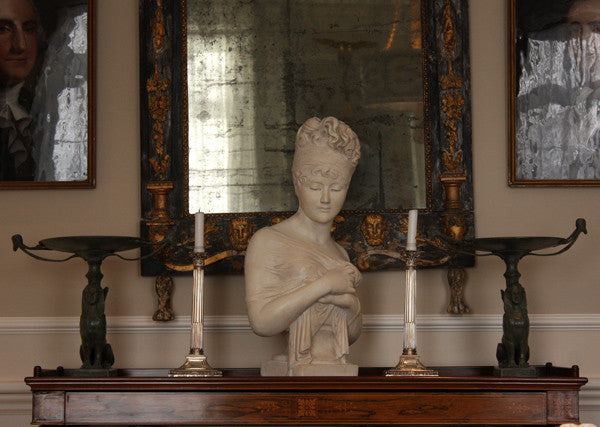 photo of plaster cast sculpture bust of Madame Recamier with high up-do and ribbon over forehead and hands crossed over chest covered in cloth on a dark wood table with bronze decorations, candlesticks, and bronze framed mirror and two pictures