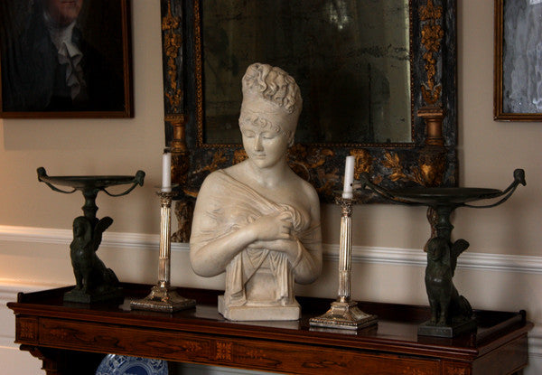 photo of plaster cast sculpture bust of Madame Recamier with high up-do and ribbon over forehead and hands crossed over chest covered in cloth on a dark wood table with bronze decorations, candlesticks, and bronze framed mirror and two pictures