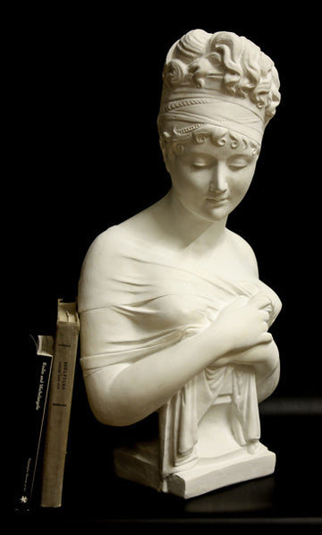 photo with black background of plaster cast sculpture bust of Madame Recamier with high up-do and ribbon over forehead and hands crossed over chest covered in cloth, and two books leaning against her