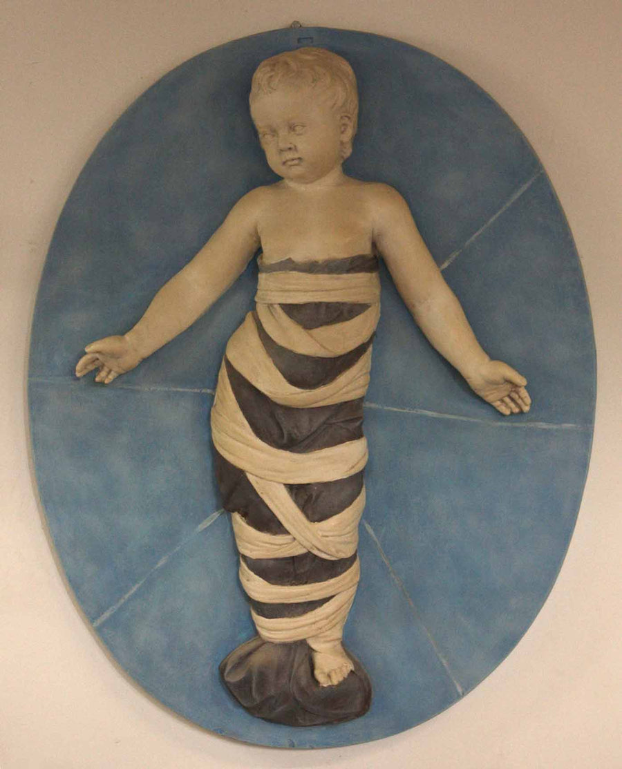 photo of plaster cast relief of child with mummy-like wrappings on a blue oval background on a tan-colored wall
