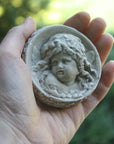 Photo of small round plaster sculpture medallion of head of an infant held by a light-skinned female hand with a blurry green background