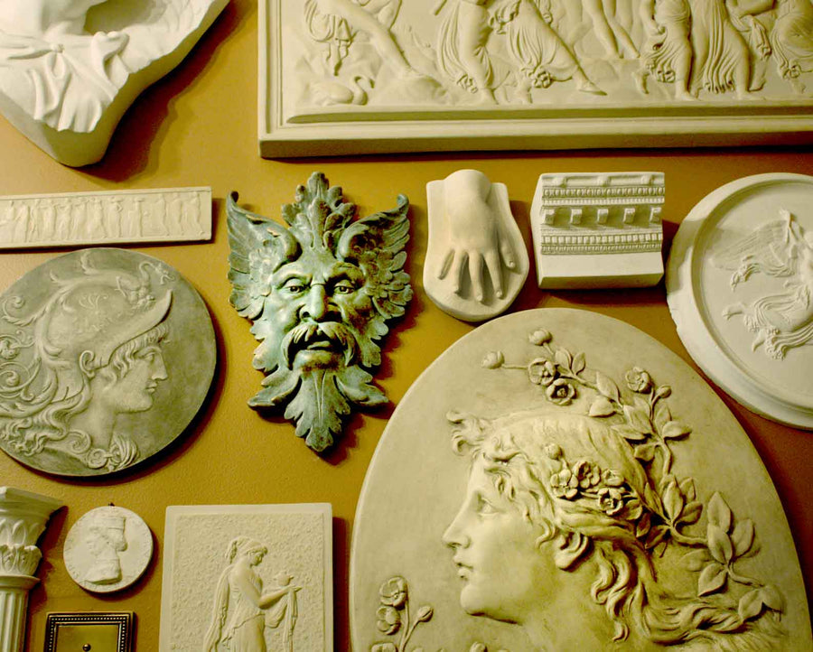 photo of plaster cast sculpture reliefs on orange-brown wall, including verdigris-colored relief of moustached man's face made of leaves