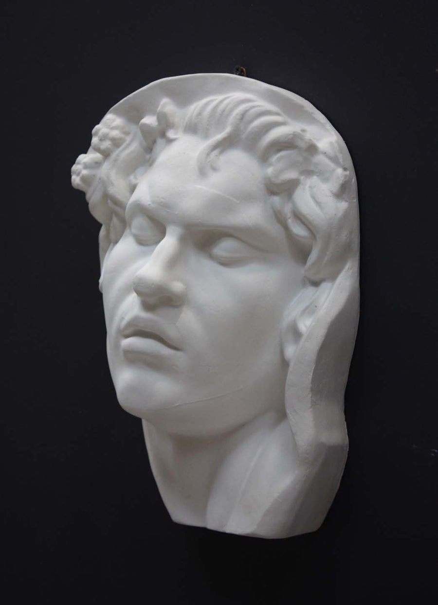 photo of plaster cast of sculpture of face of Sleeping Faun with two flowers in curly hair on dark gray background
