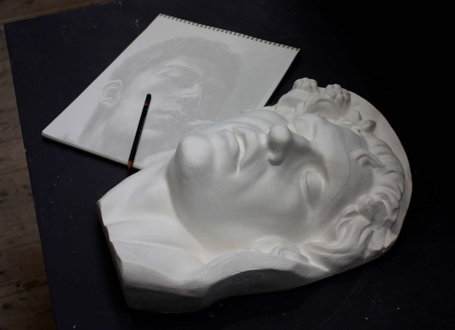 photo of plaster cast of sculpture of face of Sleeping Faun with two flowers in curly hair on dark gray surface with sketchbook and pencil beside it