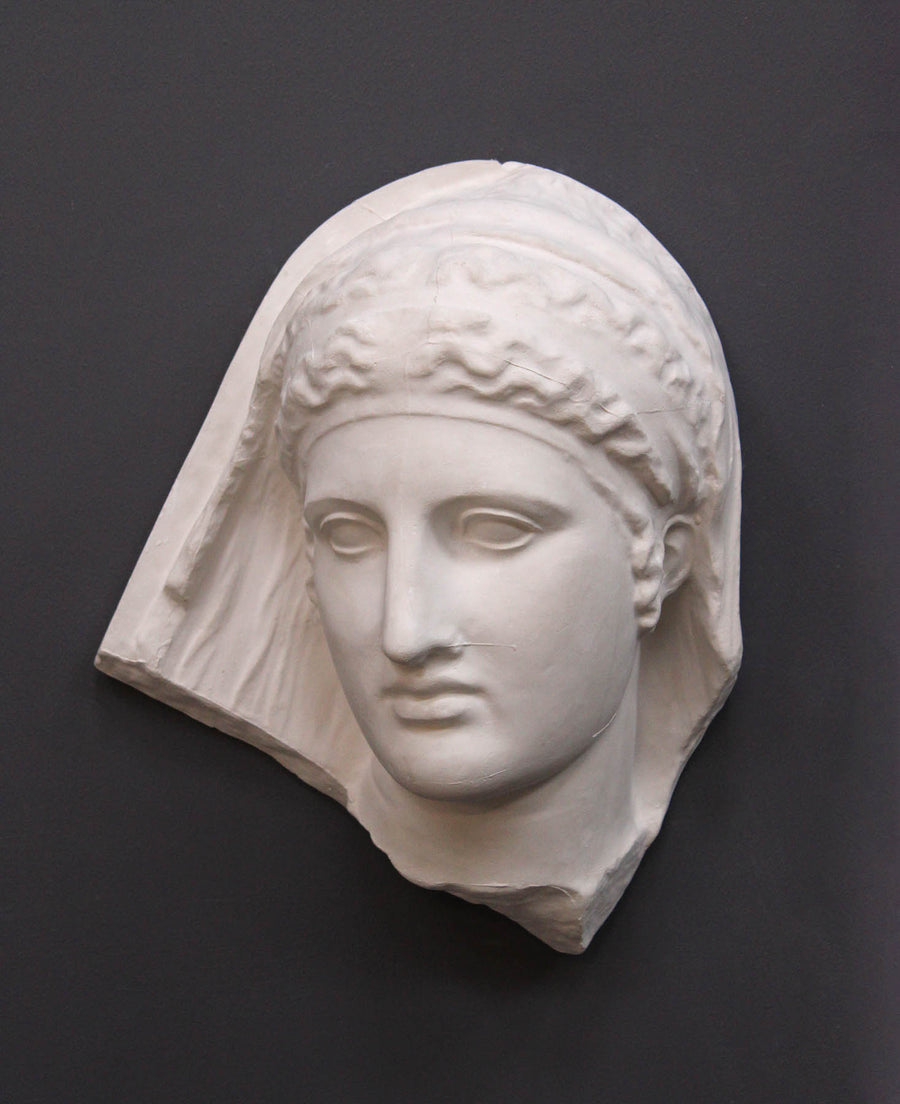 photo of white plaster cast of ancient relief sculpture of female head with headpiece and covering against dark gray background