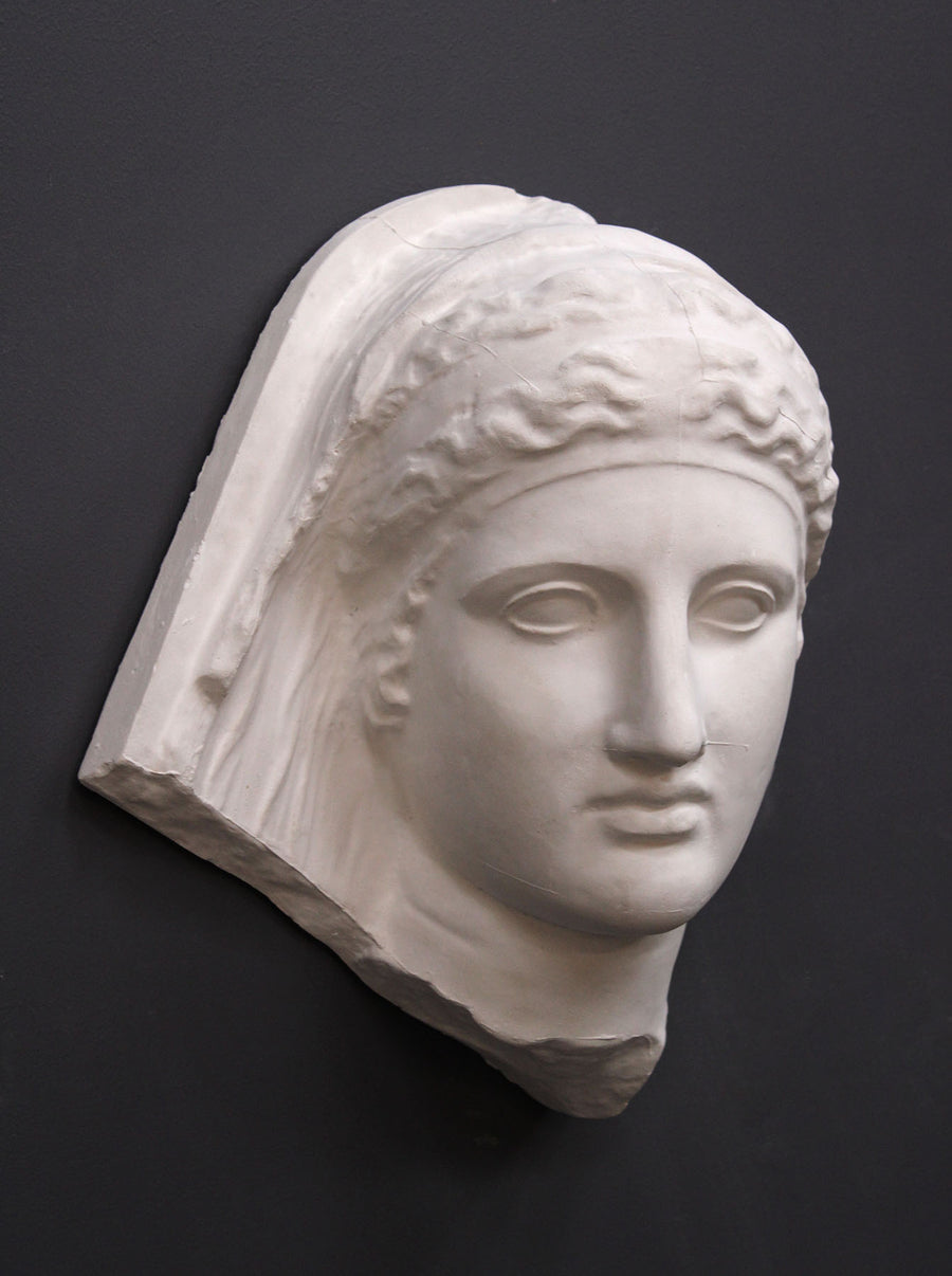 photo of white plaster cast of ancient relief sculpture of female head with headpiece and covering against dark gray background