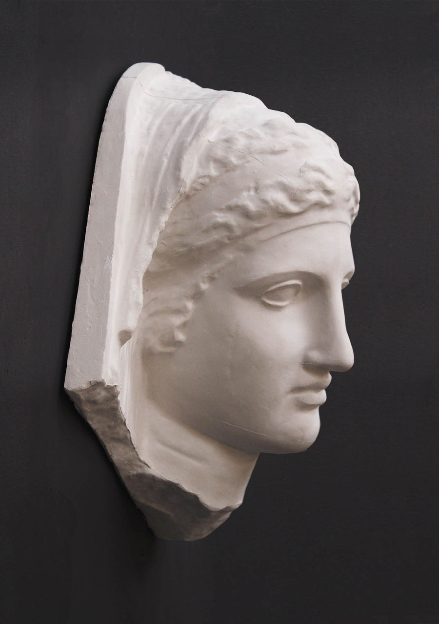 photo of white plaster cast of ancient relief sculpture of female head with headpiece and covering against dark gray background