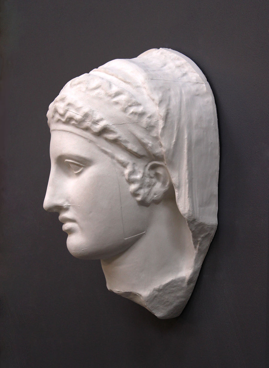 photo of white plaster cast of ancient relief sculpture of female head with headpiece and covering against dark gray background