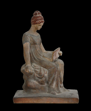 Photo of plaster cast sculpture of female in green-brown tinted robes seated with opened book against black background