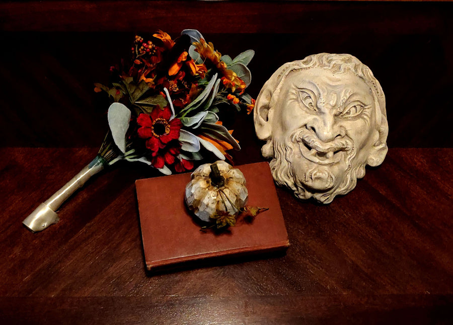 photo of plaster cast sculpture of grotesque faun face on a dark table next to fall flowers, a pumpkin, and a red book