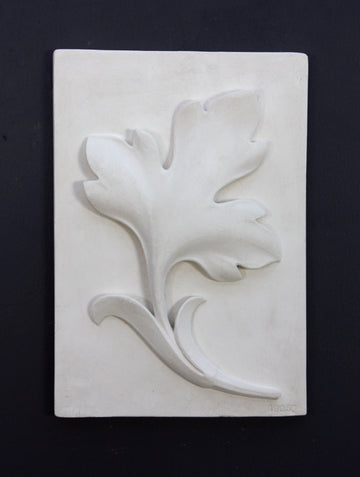 Photo of a plaster cast of a rectangle slab with a study of an acanthus leaf poited upward on the front, on a black background