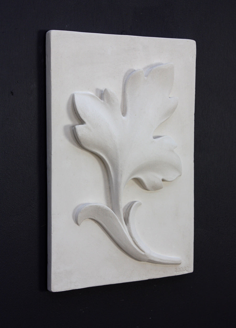 Photo of a plaster cast of a rectangle slab with a study of an acanthus leaf poited upward on the front, the whole object turned to the face the right to be seen at a 3/4 view, on a black background