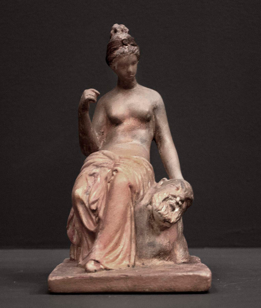photo of plaster cast sculpture of female half nude with pink robes seated and holding a mask, set against gray background