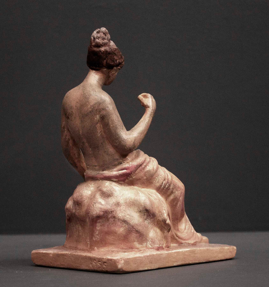 photo of plaster cast sculpture of female half nude with pink robes seated and holding a mask, set against gray background