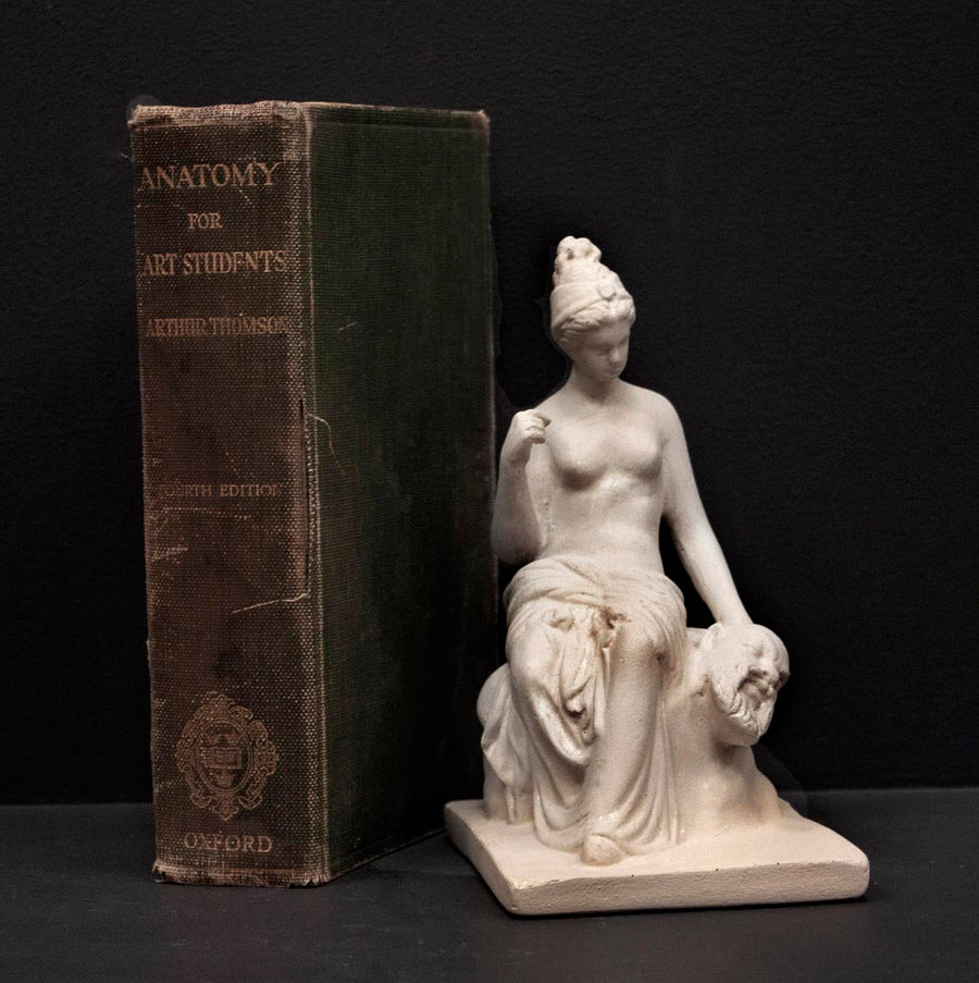 photo of white plaster cast sculpture of female half nude seated and holding a mask, set beside green book and against gray background