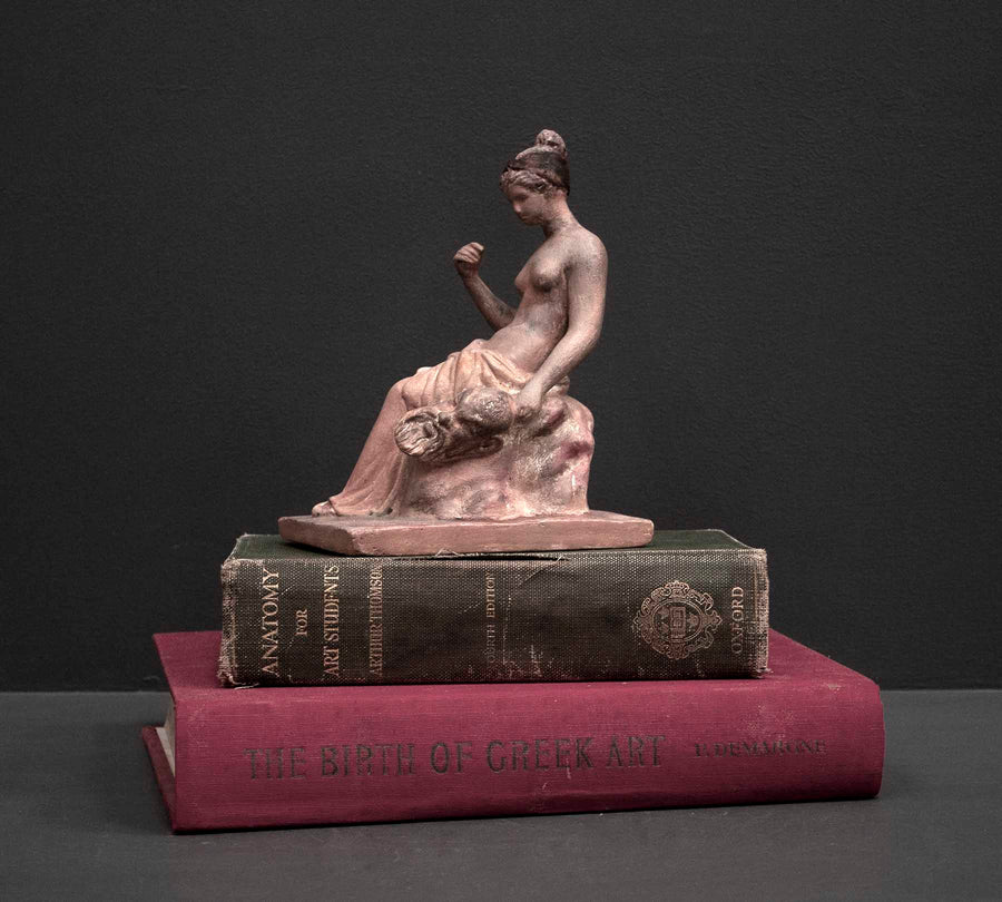 photo of plaster cast sculpture of female half nude with pink robes seated and holding a mask, set atop a green book and a red book against gray background
